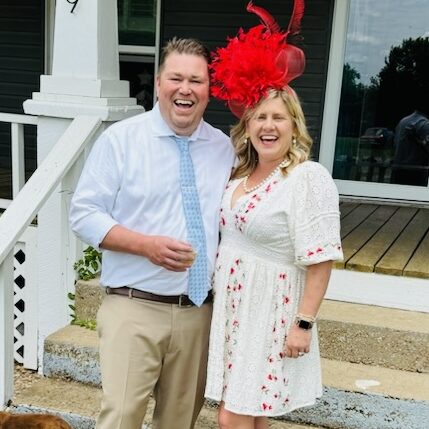 Tara and her husband Jim dressed up for derby day. Jim is wearing a blue shirt and tie while Tara wears a wacky red derby hat and a matching red and white dress.
